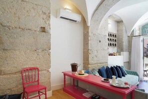 Una habitación con pared de piedra, una silla roja, una mesa con una cafetera y una cama con almohadas.