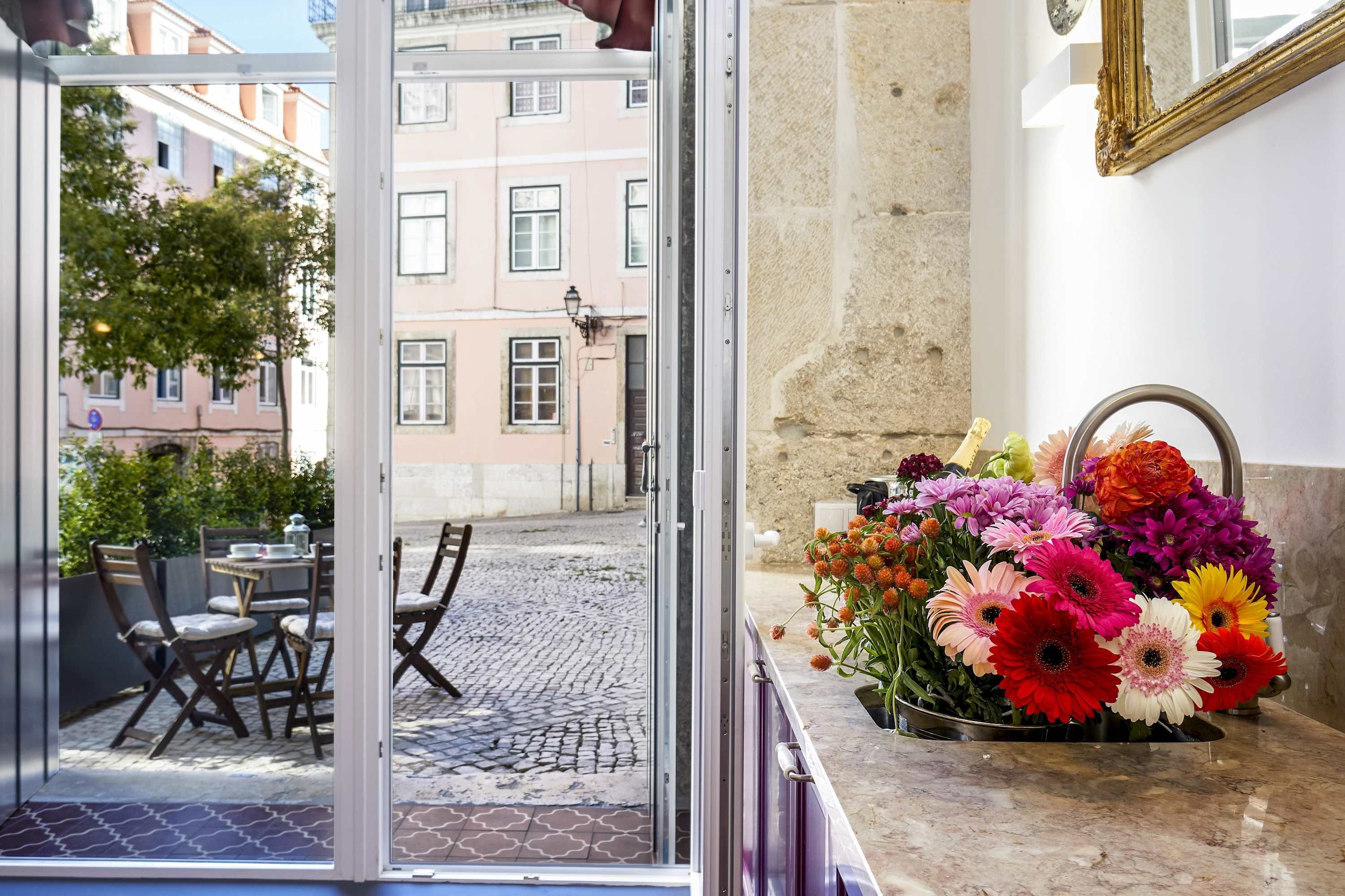 Ein Hotelzimmer mit Blick auf eine Kopfsteinpflasterstraße und umliegende Gebäude. Vor dem Zimmer steht ein Tisch mit Stühlen, auf der Anrichte steht eine Blumenvase.