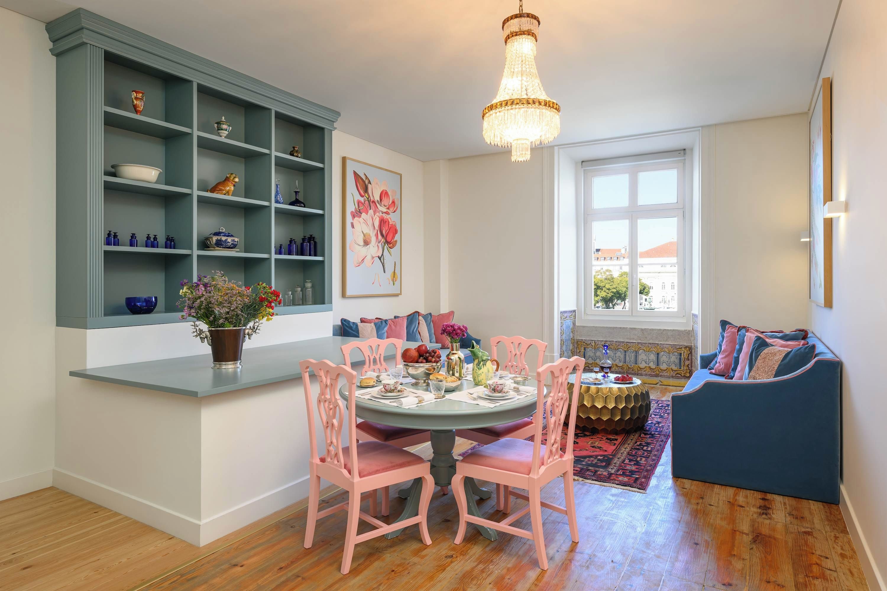Uma sala de jantar com uma mesa redonda, cadeiras cor-de-rosa e um armário azul-esverdeado com prateleiras.