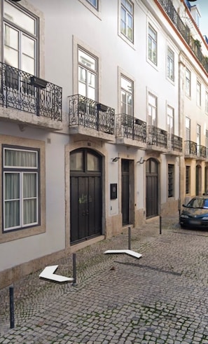 A row of white buildings with black doors and windows, a cobblestone street, and a car parked on the right. - Lisbon Duplex Apartment with 2 Bedrooms & Balcony (Lisbon)