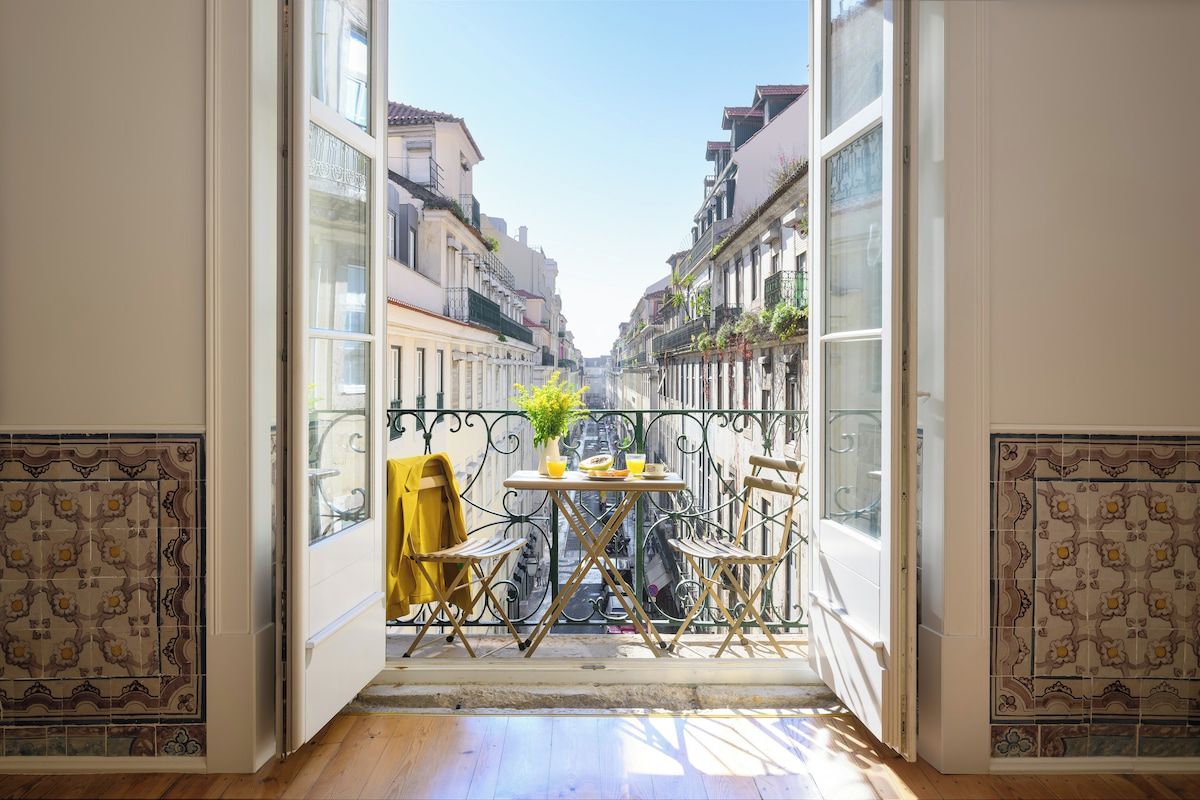 Un balcón abierto con una mesa y sillas, una planta en maceta y vista a una calle estrecha con edificios.