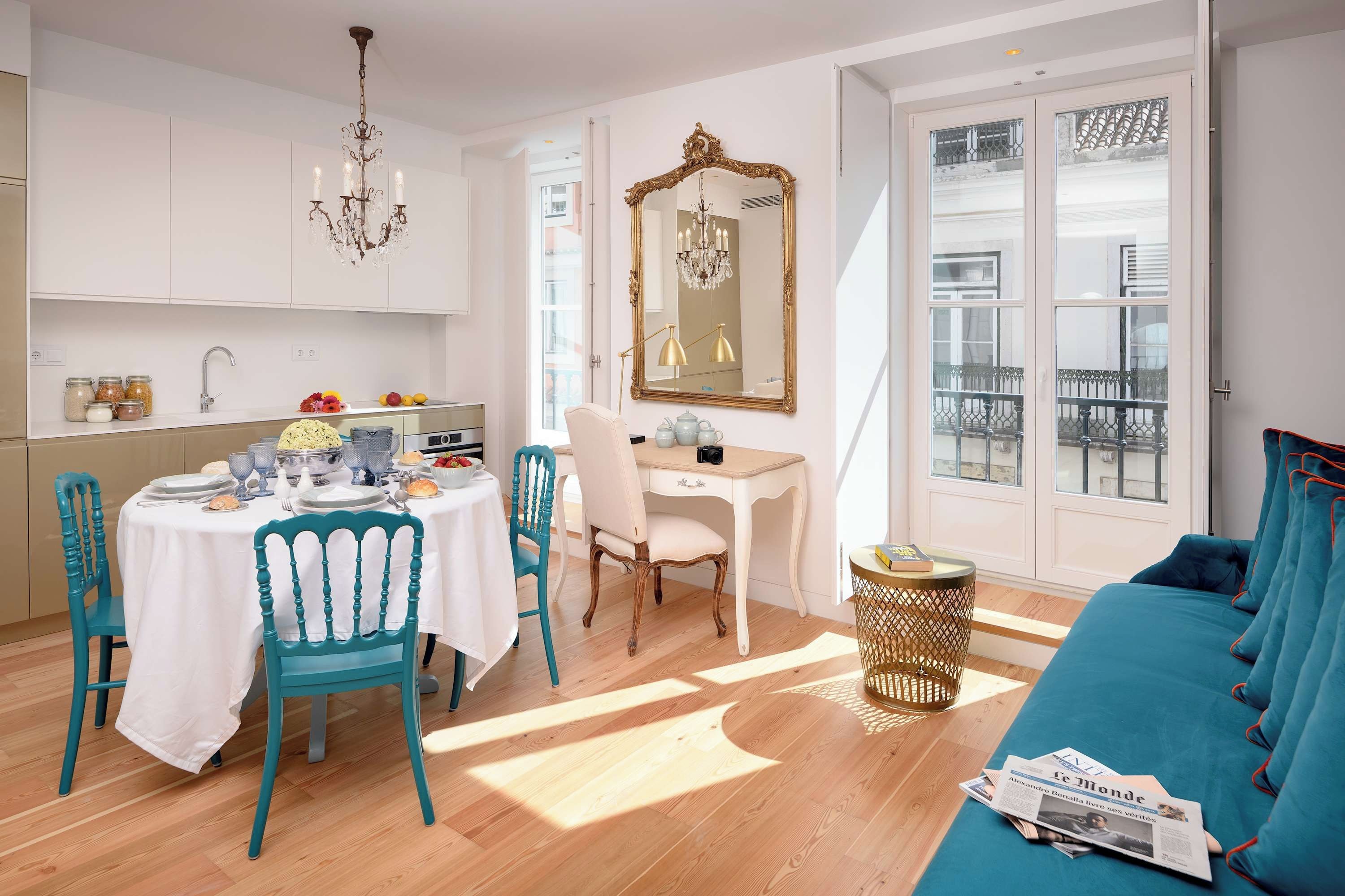 Una habitación bien iluminada con una mesa de comedor para cuatro personas, un escritorio con silla, un espejo grande y un sofá color turquesa con un periódico.