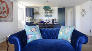 En stue med en blå sofa, to blomstrede puder, et spisebord og et køkken i baggrunden.
