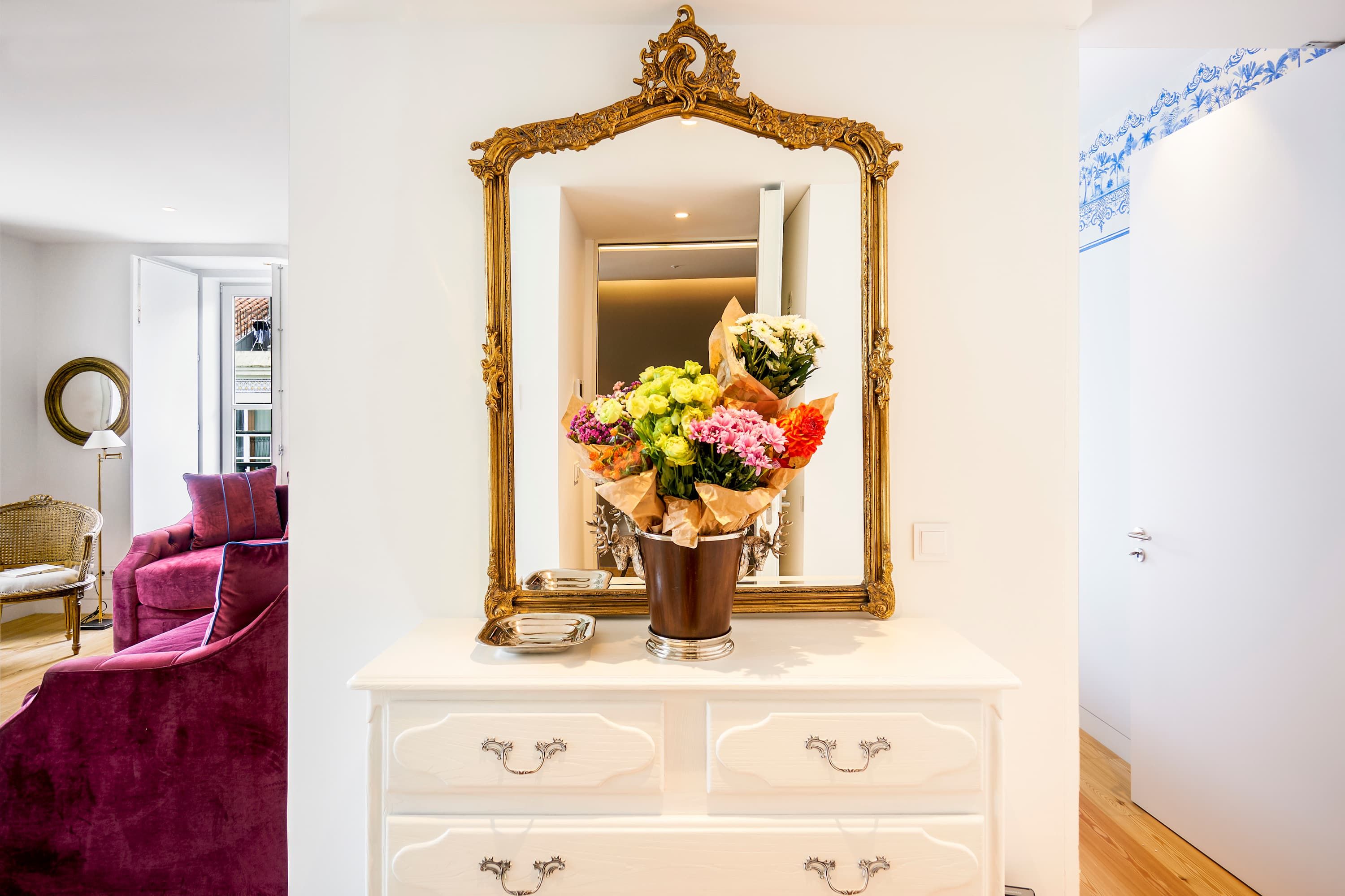 Quarto com cômoda branca, espelho com moldura dourada elaborada e um vaso de flores.