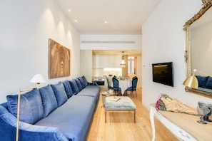 Una sala de estar moderna con un sofá azul, un comedor con mesa y sillas, una cocina al fondo y un espejo grande en la pared.