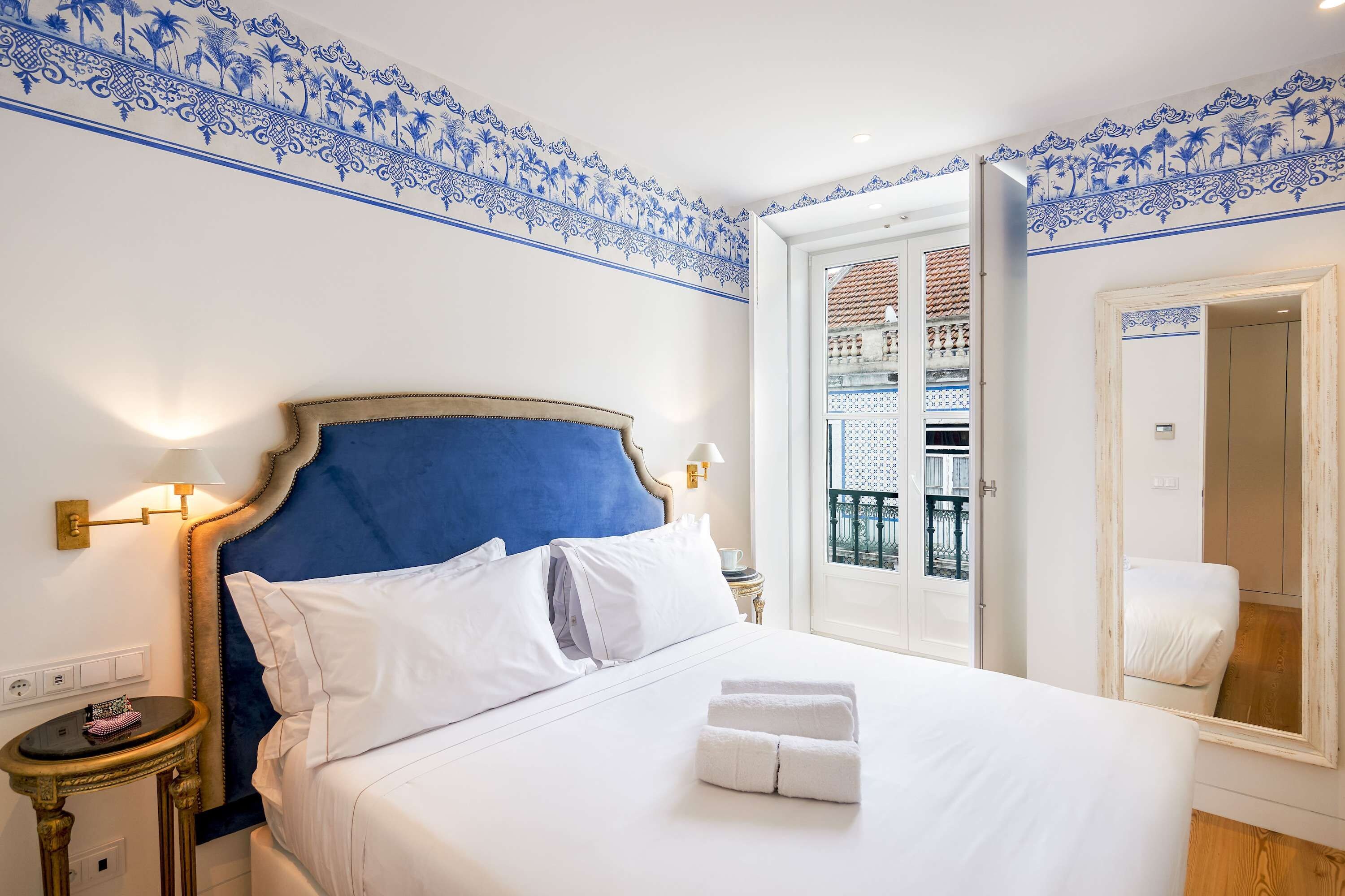 Quarto de hotel com uma cama grande, uma mesa de cabeceira, uma luminária e vista para a varanda externa.