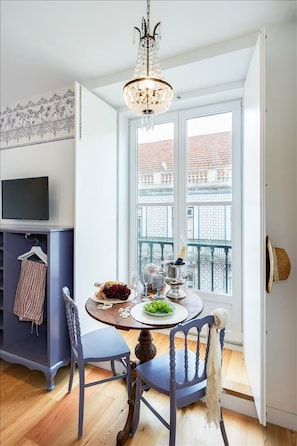 Un comedor con candelabro, una mesa servida con platos, vasos y una botella, y vistas a edificios a través de una ventana.