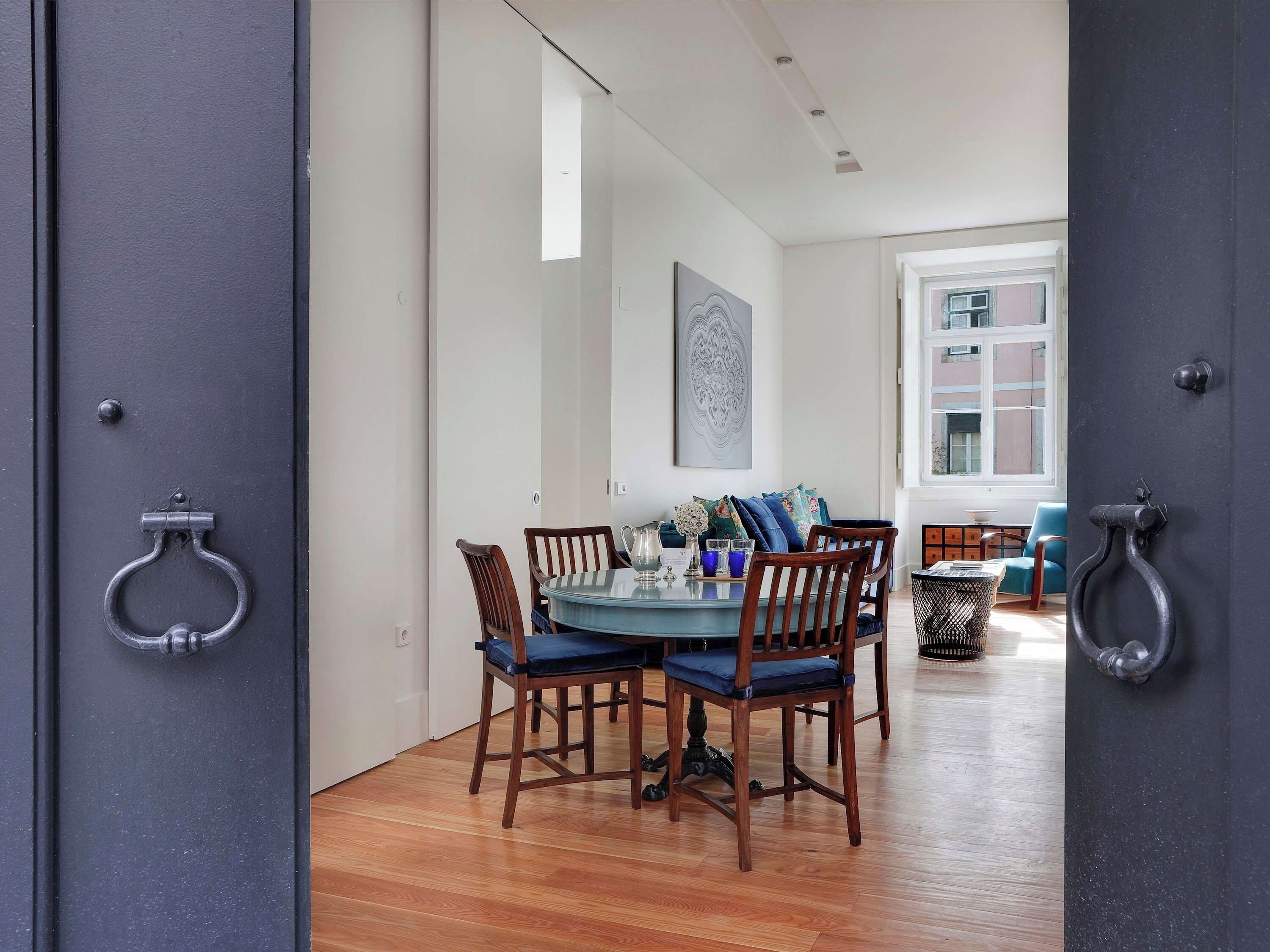 A dining area with a round table and chairs, a window with a view of buildings, and a door with a metal handle.