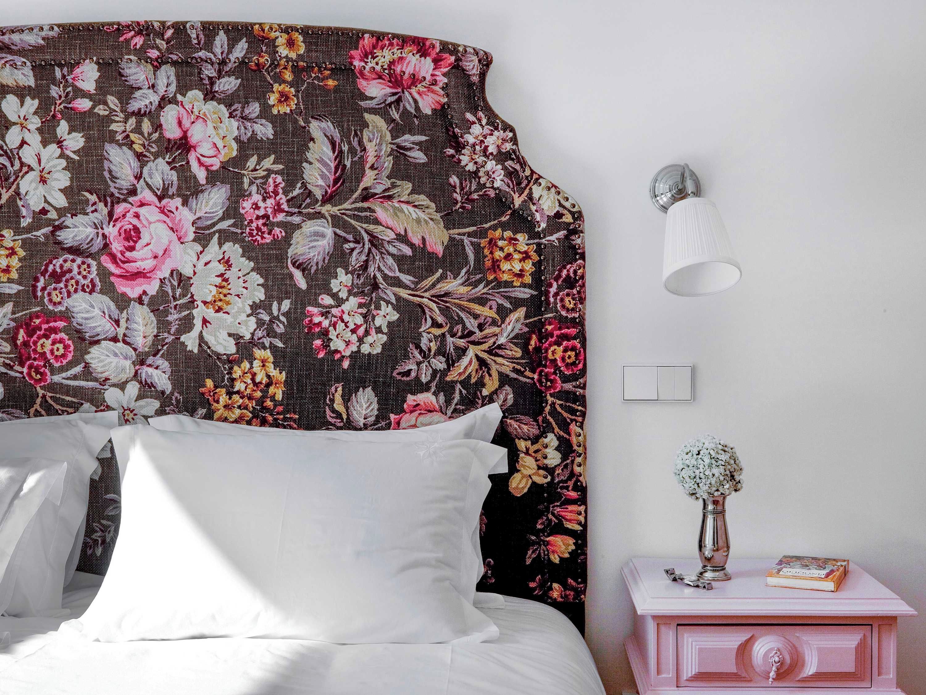 A bedroom with a floral headboard, a bedside lamp, a nightstand with a vase and book, and a white bed.