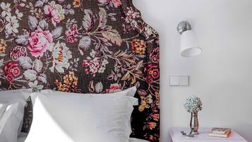 A bedroom with a floral headboard, a bedside lamp, a nightstand with a vase and book, and a white bed.