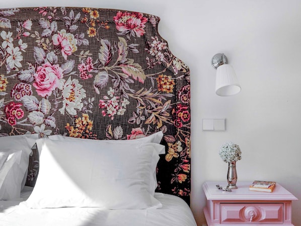 A bedroom with a floral headboard, a bedside lamp, a nightstand with a vase and book, and a white bed. - Historic Lisbon Apartment Near Chiado (Lisbon)