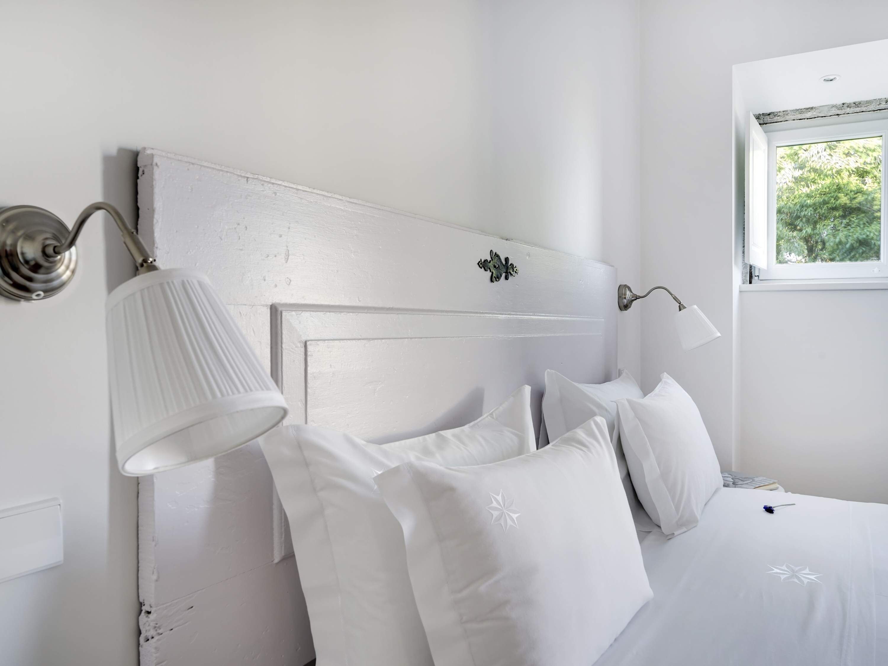 Ein helles Schlafzimmer mit einem Bett, Kissen, einem Nachttisch, einer Lampe und einem Fenster mit Blick auf Grünfläche.