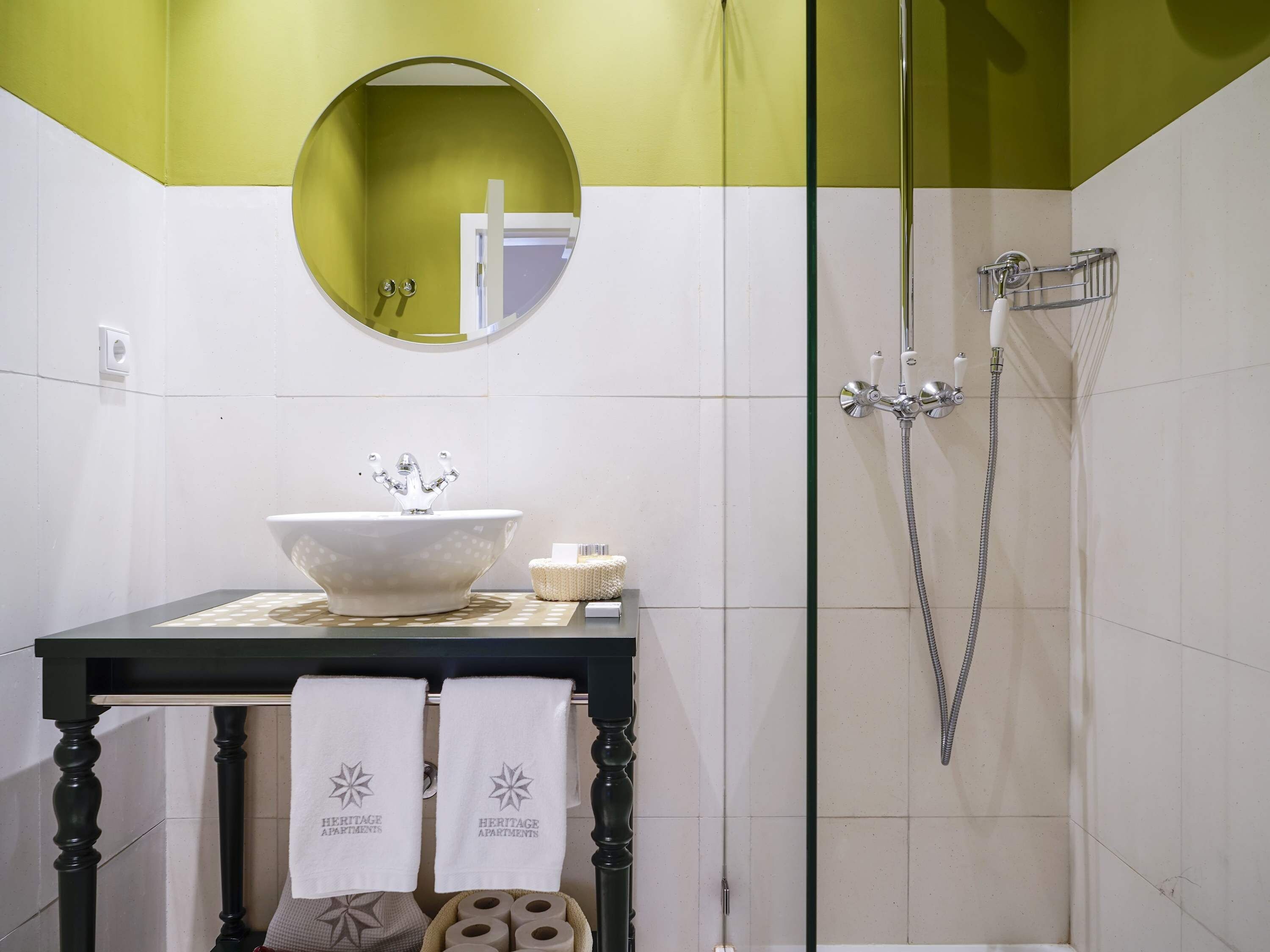 Een badkamer met een ronde spiegel, een wastafel met een decoratieve wasbak, een douche met een handdouche en handdoeken aan een rek.