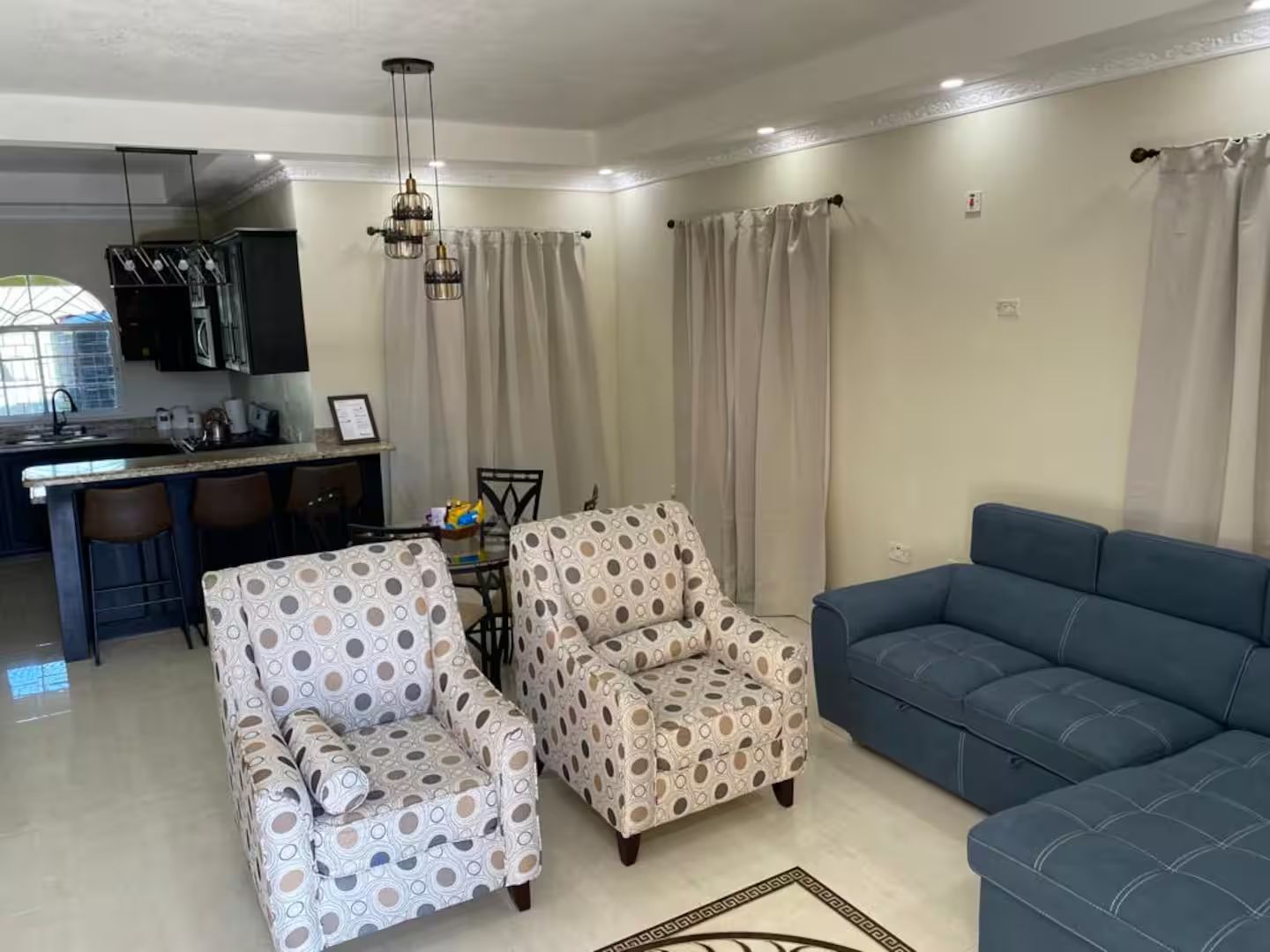 A living room with a sofa, armchairs, a dining table, and a kitchen area with a window.
