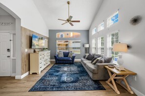 A living room with a ceiling fan, a blue sofa, a grey sofa, a wooden coffee table, and a patterned rug. - Emerald Island Corner Townhome  2 King Suites (Kissimmee)