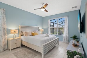 Ein Schlafzimmer mit Bett, Nachttischen, Deckenventilator, Fenster mit Jalousien und einem an der Wand montierten Fernseher.