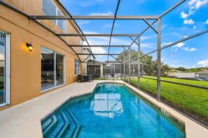 Ein Schwimmbecken mit überdachter Terrasse, ein Haus mit großen Fenstern und ein klarer Himmel.
