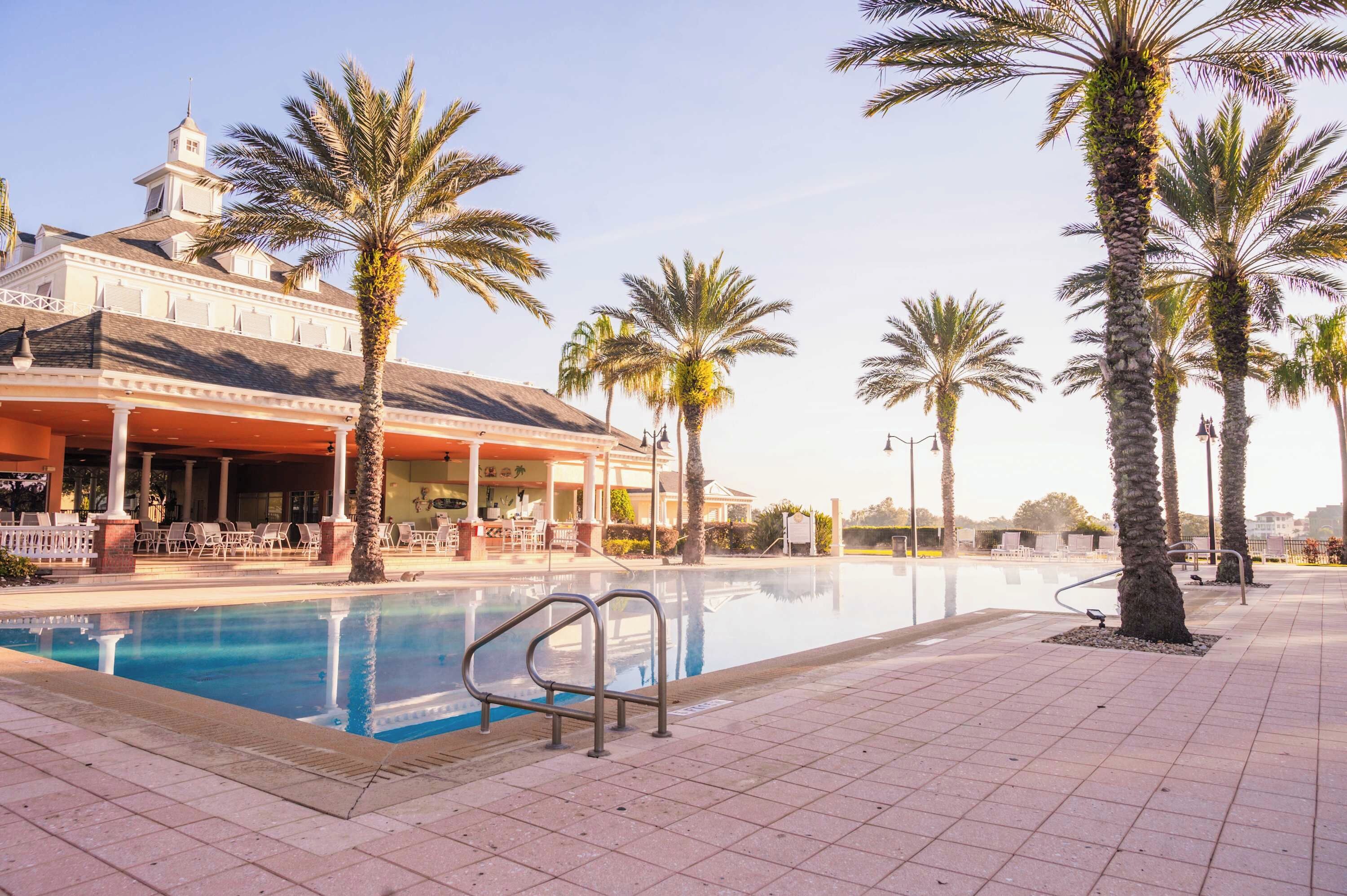 Zona de piscina con palmeras, un área de descanso cubierta y un edificio con torre.