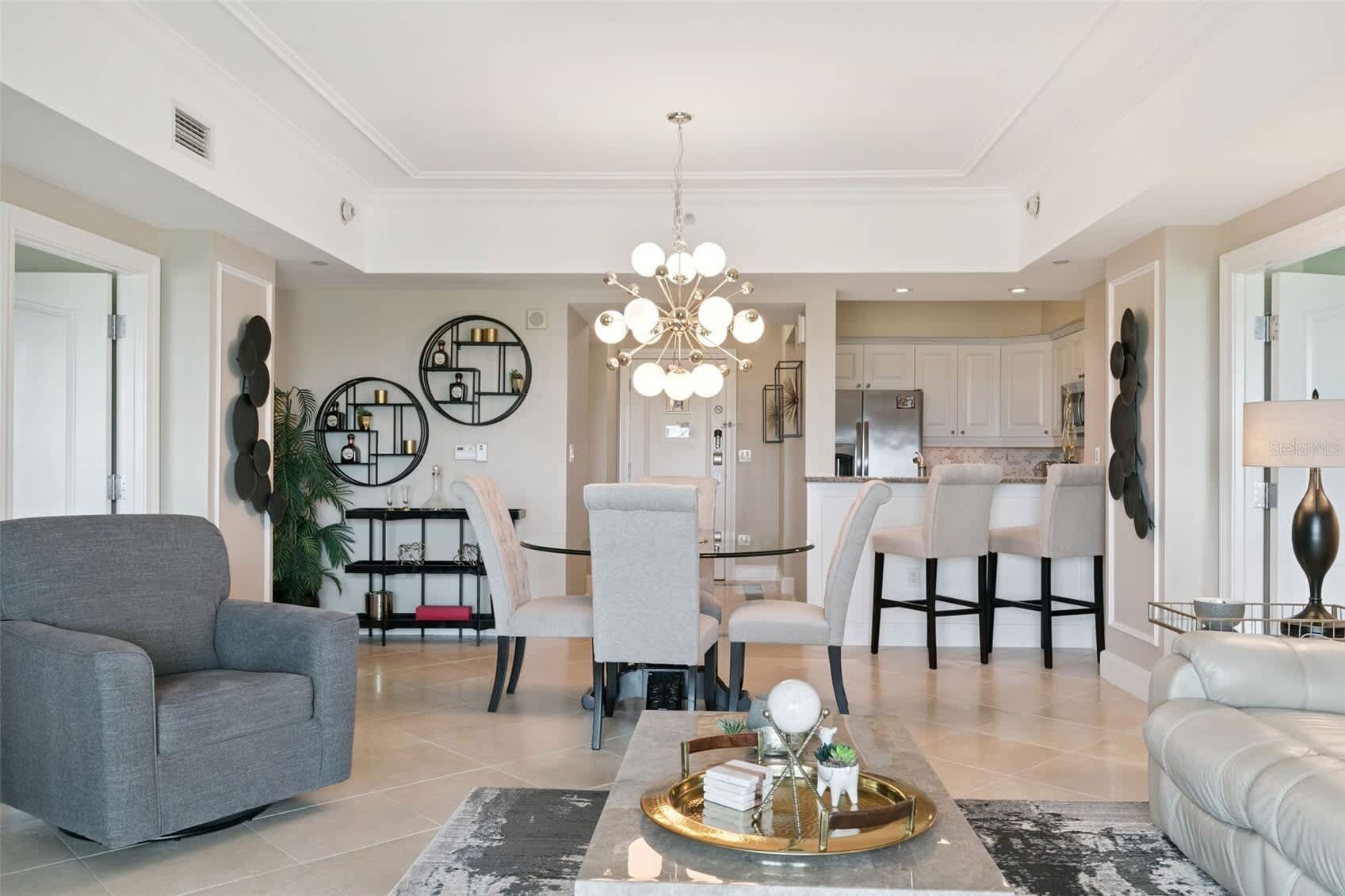 Una sala de estar moderna con un sofá gris, un comedor con una araña y una cocina al fondo.