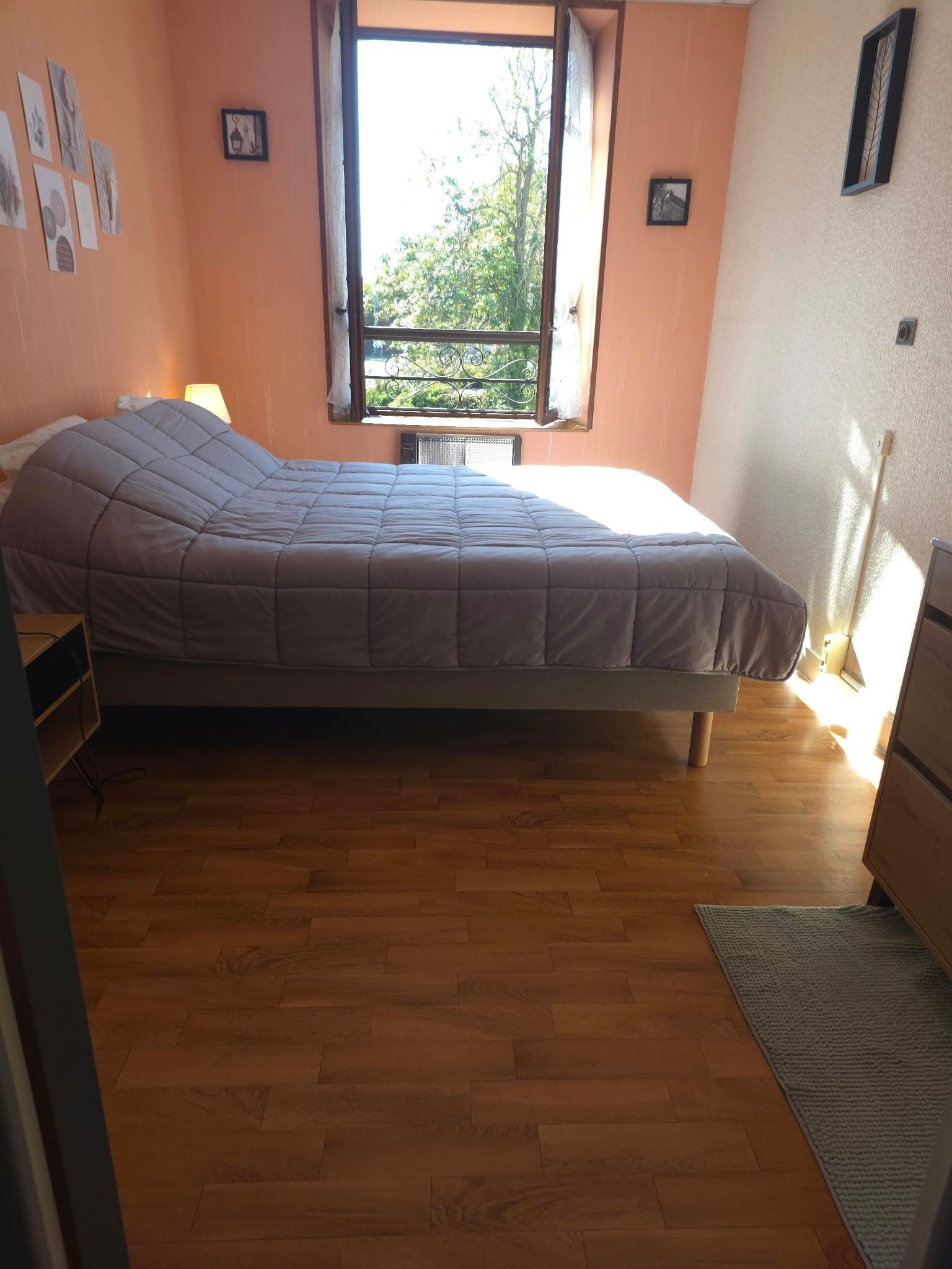 Een eenpersoonsbed in een kamer met houten vloeren, een raam met uitzicht op bomen en een muur met ingelijste foto's.