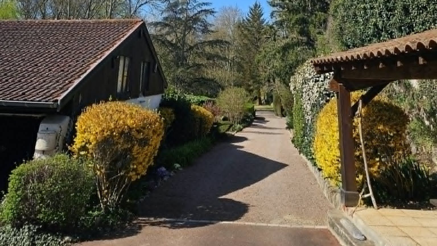 Un chemin menant à un bâtiment dont le toit est recouvert de tuiles, entouré d’arbustes aux fleurs jaunes et de verdure.
