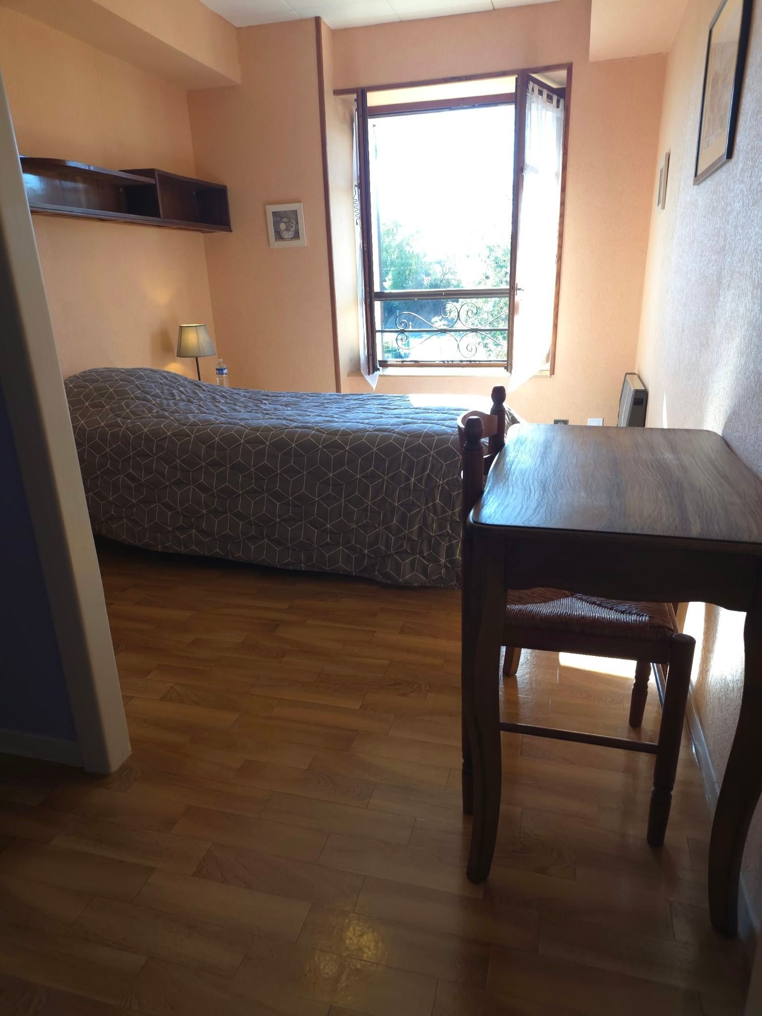 Un dormitorio con cama, mesa, silla, ventana y un cuadro en la pared.