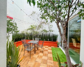 Eine Terrasse mit Holzboden, einem Metalltisch und Stühlen. Umgeben von Bäumen und verschiedenen Pflanzen.