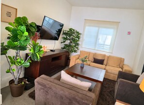 Ein Wohnzimmer mit Flachbildfernseher, einer braunen Unterhaltungsecke, einem beigen Sofa, einem Couchtisch und zwei Blumentöpfen.