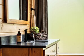 Ein Badezimmer-Waschtisch mit Holzschrank, schwarzer Arbeitsplatte, einer Handseifenflasche, einem schwarzen Wasserhahn und einem gewebten Korb mit Pflanze.