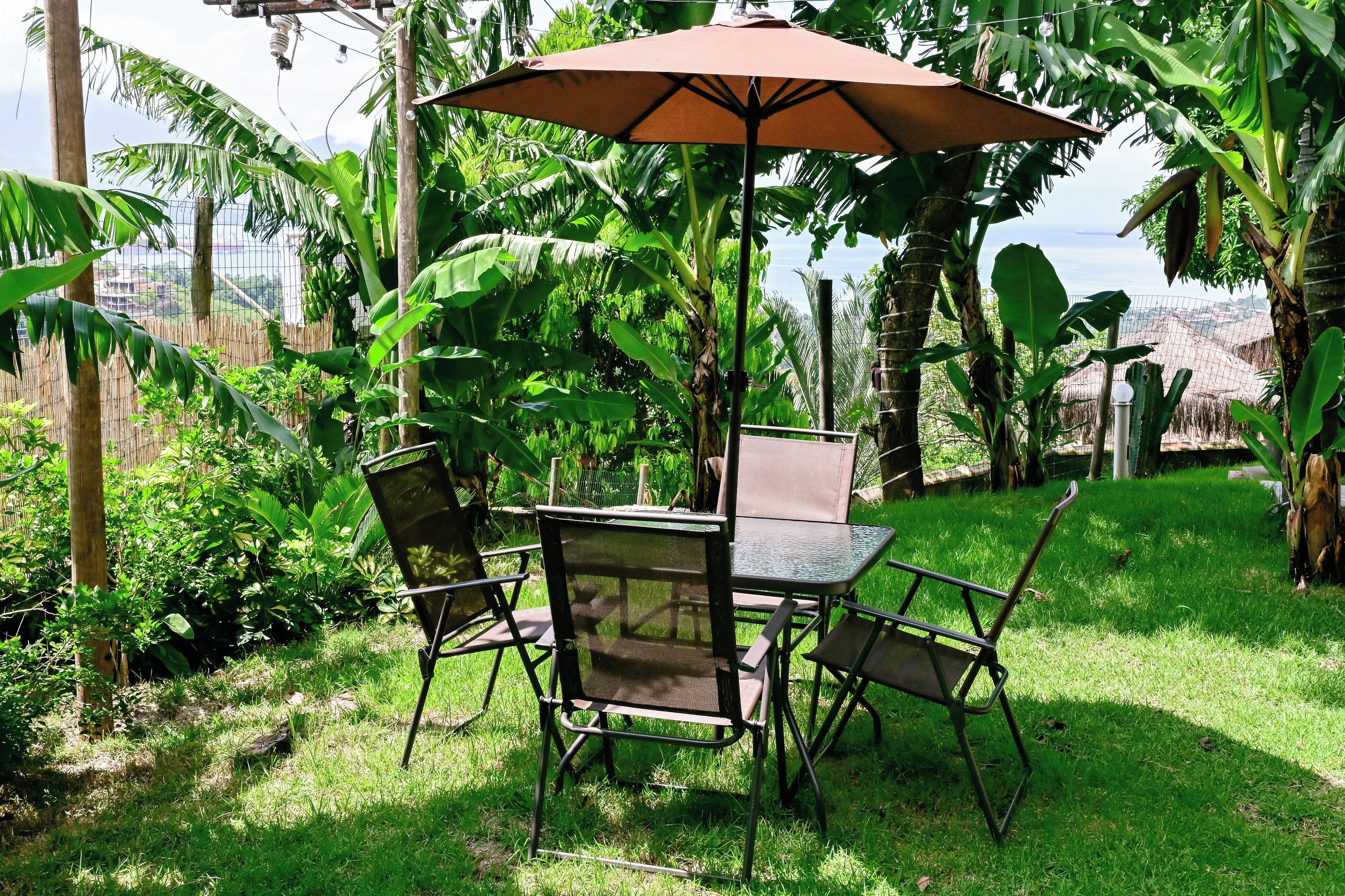 An outdoor seating area with a table and chairs under a large umbrella, surrounded by lush greenery.