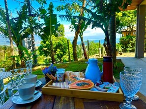 A breakfast tray with food and drinks on a wooden table outdoors with a view of the sea and greenery. - Studios Ilhabela (Ilhabela)