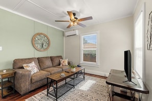 A living room with a brown sofa, a coffee table, a TV stand, a ceiling fan, and a window with blinds. - The Nook on 4th Street (Prescott)