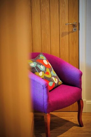 Un fauteuil violet agrémenté d’un coussin aux motifs colorés représentant des oiseaux, placé devant une porte en bois.