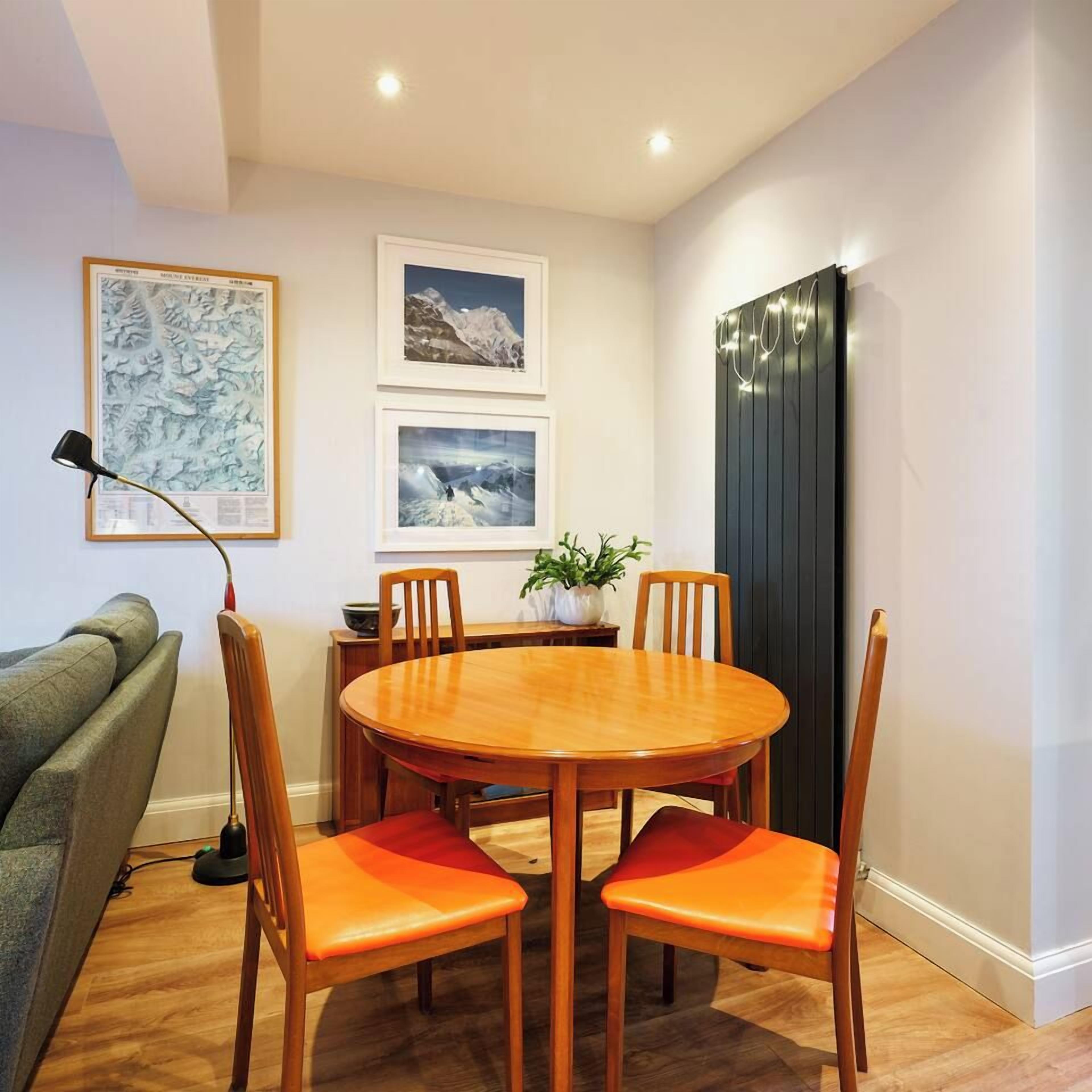 A dining area with a round wooden table and four chairs with orange cushions, a grey sofa, and a floor lamp.