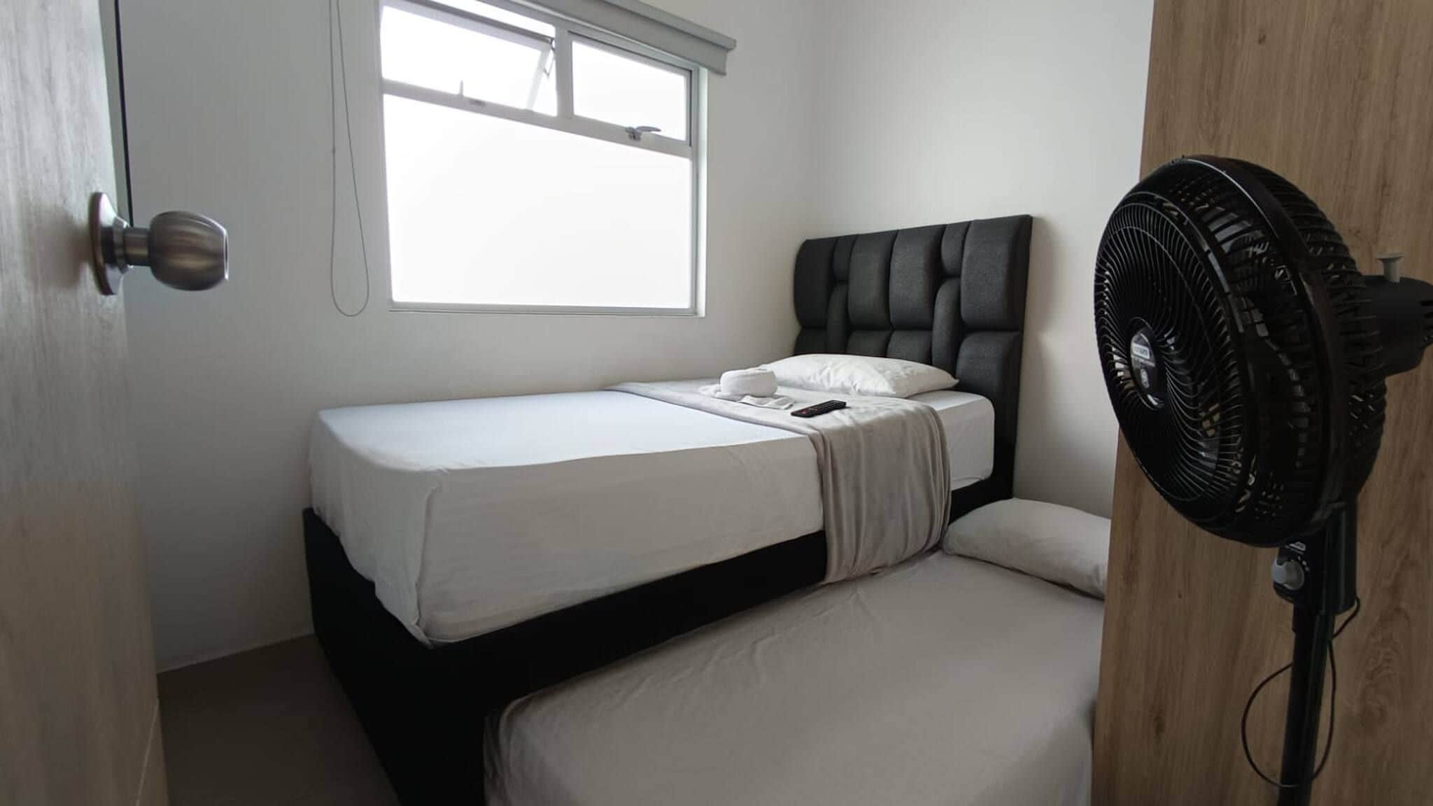 A single bed with a white mattress and a dark headboard in a room with a window and a standing fan.