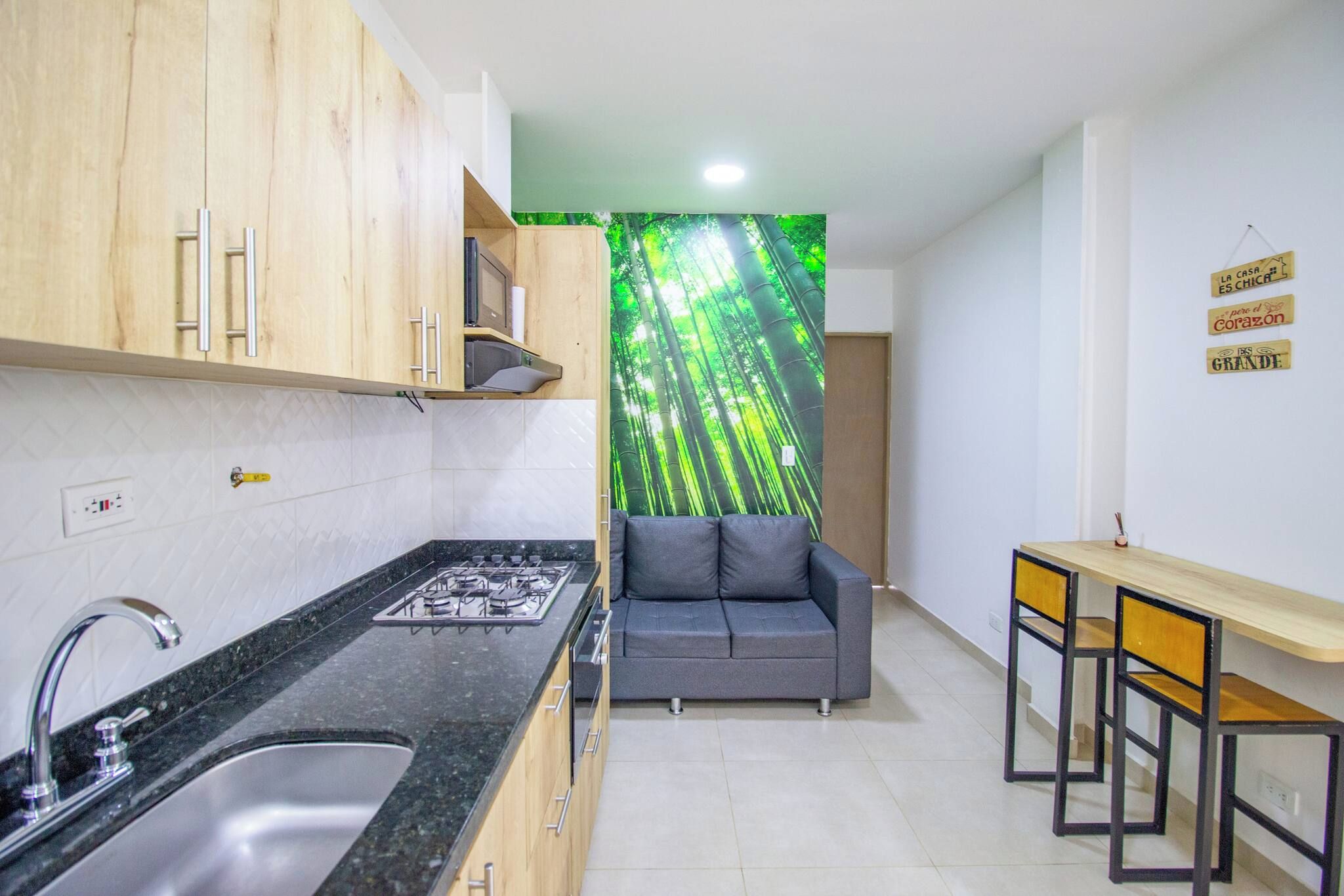 Una cocina funcional con lavamanos, estufa y armarios de madera. Un sofá gris y una pequeña mesa de comedor con sillas se observan al fondo.