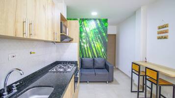 Una cocina funcional con lavamanos, estufa y armarios de madera. Un sofá gris y una pequeña mesa de comedor con sillas se observan al fondo.