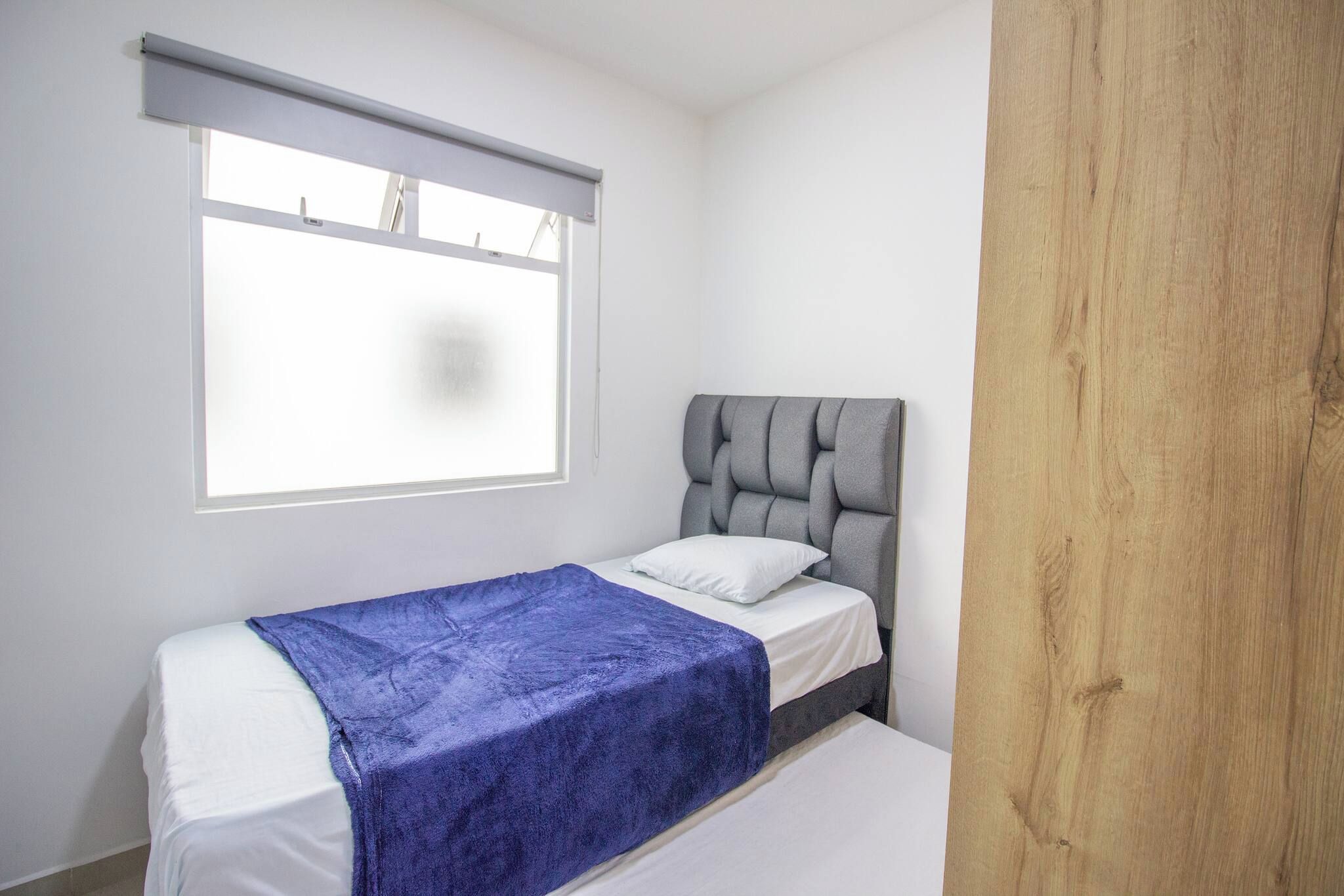 Una cama individual con cabecera gris y manta azul, una ventana con persiana y un panel de madera en la pared.