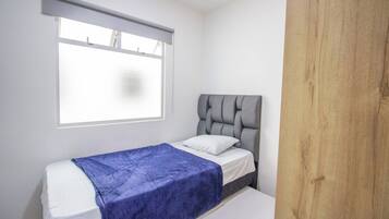 Una cama individual con cabecera gris y manta azul, una ventana con persiana y un panel de madera en la pared.