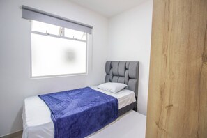 Una cama individual con cabecera gris y manta azul, una ventana con persiana y un panel de madera en la pared.