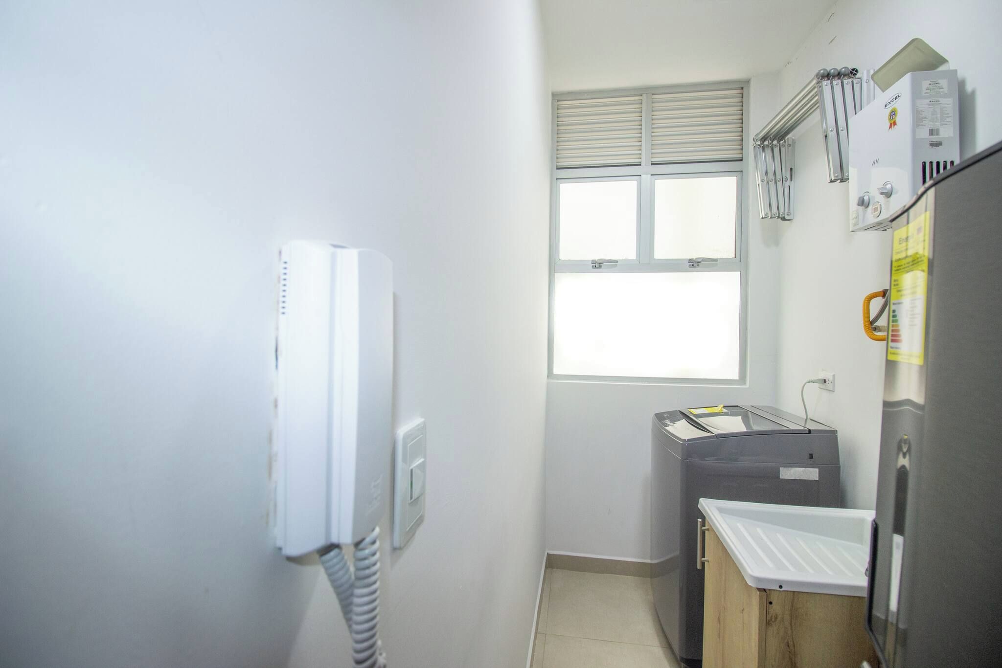 Una habitación pequeña y funcional con ventana, un calentador blanco, un electrodoméstico gris y una mesita con superficie blanca.