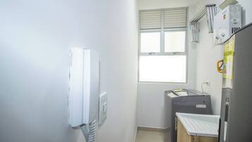 Una habitación pequeña y funcional con ventana, un calentador blanco, un electrodoméstico gris y una mesita con superficie blanca.