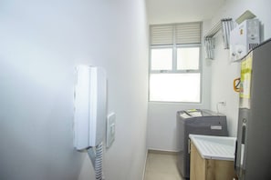 Una habitación pequeña y funcional con ventana, un calentador blanco, un electrodoméstico gris y una mesita con superficie blanca.