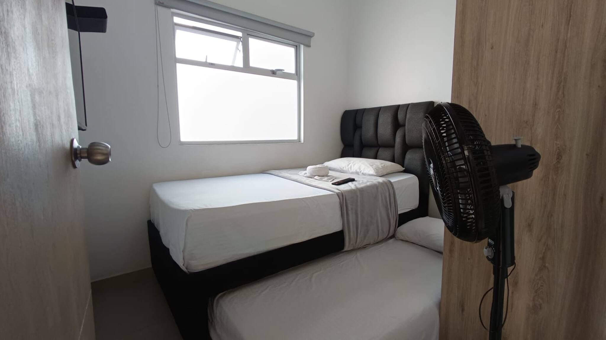 Una cama individual con colchón blanco y cabecera gris en una habitación con ventana y puerta.