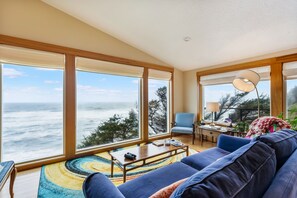 A spacious living room with a large window overlooking the ocean, a blue sofa, a coffee table, and a desk with a lamp. - Sundown Cliffs- Premium Coastal Getaway with Breathtaking Ocean Views & Sunsets (Cloverdale)