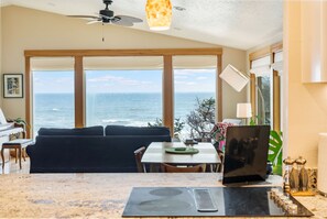 A room with a large window overlooking the ocean, a ceiling fan, a dining table with chairs, and a black countertop with a laptop. - Sundown Cliffs- Premium Coastal Getaway with Breathtaking Ocean Views & Sunsets (Cloverdale)
