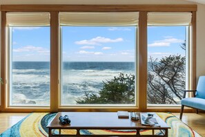 A room with a large window overlooking the ocean, a wooden table with a pair of binoculars, and a blue chair. - Sundown Cliffs- Premium Coastal Getaway with Breathtaking Ocean Views & Sunsets (Cloverdale)