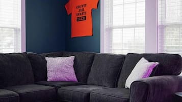 A living room with a dark blue wall, a gray sectional sofa, a round wooden coffee table, and a purple and white patterned rug.