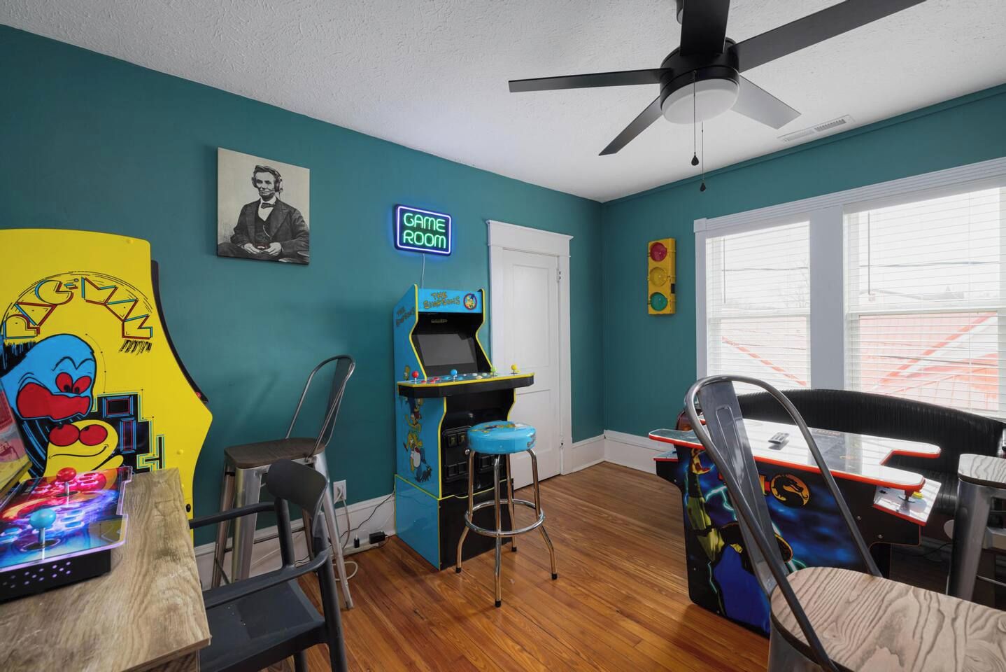 A room with teal walls, a ceiling fan, a neon sign reading 'GAME ROOM', and arcade games.