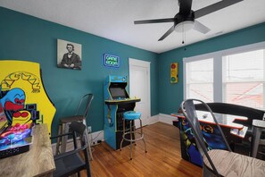 Una habitación con paredes verde azulado, un ventilador de techo, un letrero de neón que dice 'GAME ROOM' y juegos de arcade.